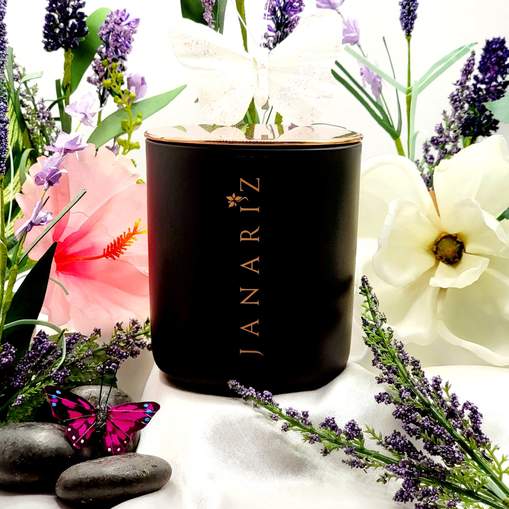 Janariz Lavender Mist luxury candle in a black jar with rose gold lettering, surrounded by lavender sprigs, white and pink flowers, black spa stones, and butterfly decorations on a white satin surface.