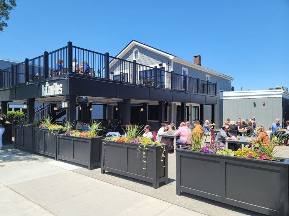 People sitting at outdoor tables under a blue sky outside a black two-story building labeled Carlton Ales. Colorful planters line the patio.