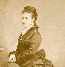 Molly Edwards - photo of a young woman in crepe early bustle dress with lace collar. Her hair is in a high bun atop her head with ornamental pins. She has a content expression.