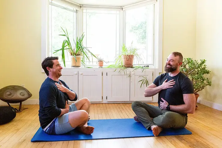 Two men sit cross-legged on a blue mat in a light-filled room with plants, smiling and touching their chests, conveying a sense of relaxation and connection.