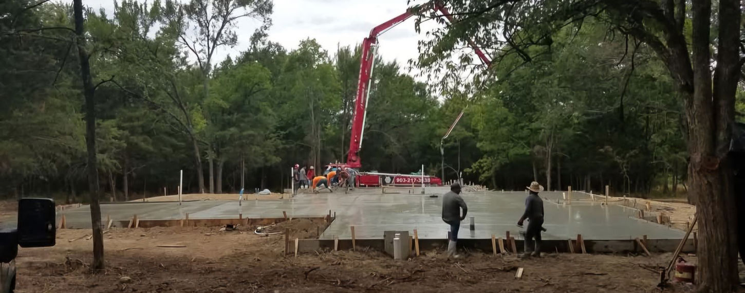Construction site with workers and a concrete pump truck pouring a large slab foundation, managed by a general contractor.