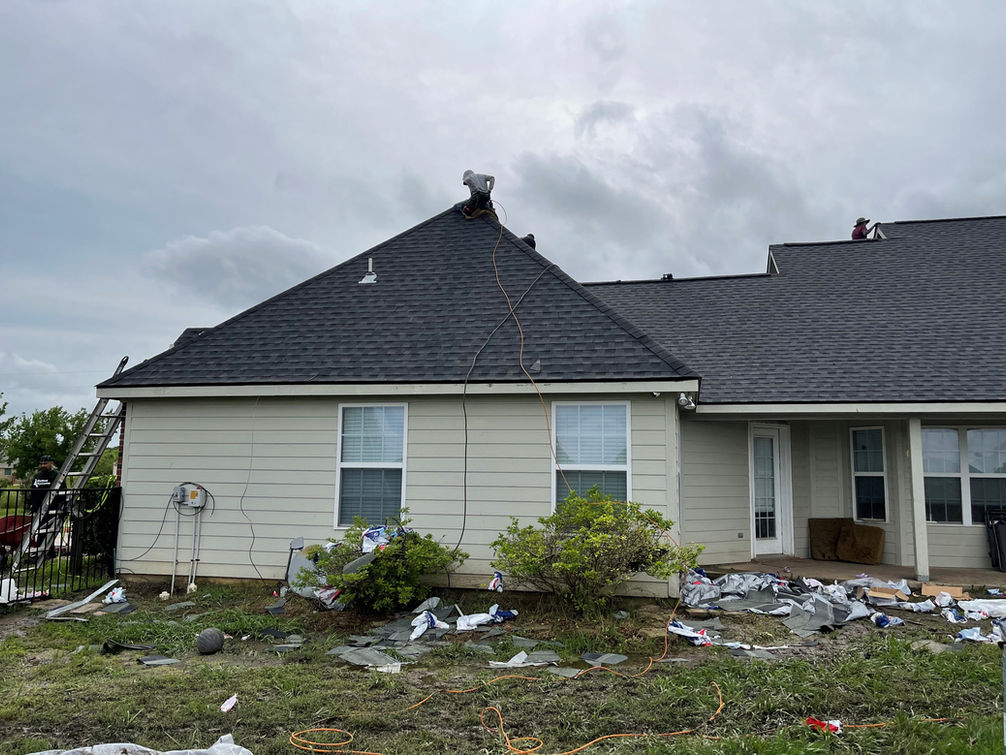 Workers repair the roof of a beige house, scattered debris on the lawn, as they tackle their latest roofing project.