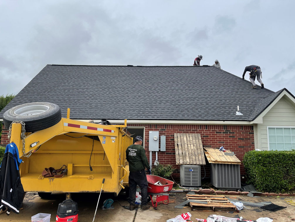Roofing workers install shingles on a house roof, while a yellow trailer is tipped over nearby, with tools and materials scattered.