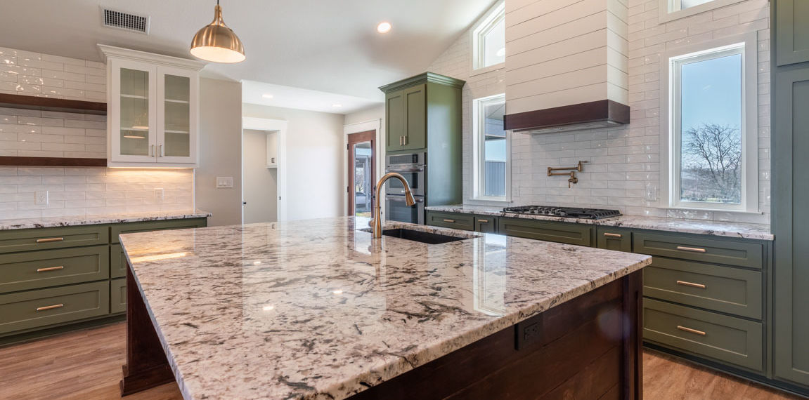 A modern kitchen with a granite island, green cabinets, wooden floors, built-in stove, pendant lighting—perfect for barndominiums.