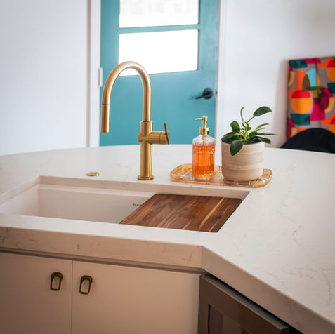 Stylish kitchen sink setup featuring a gold faucet over a white countertop, partially covered by a wooden cutting board. A decorative tray holds orange liquid soap and a small potted plant. In the background, a teal door and a colorful geometric-patterned chair add vibrant contrast—capturing AMF Construction’s blend of utility and expressive design.