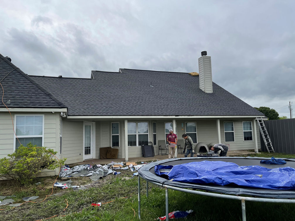 Two men stand in a backyard featuring a trampoline, patio furniture, and scattered items; the custom homes roof is dark against light siding.