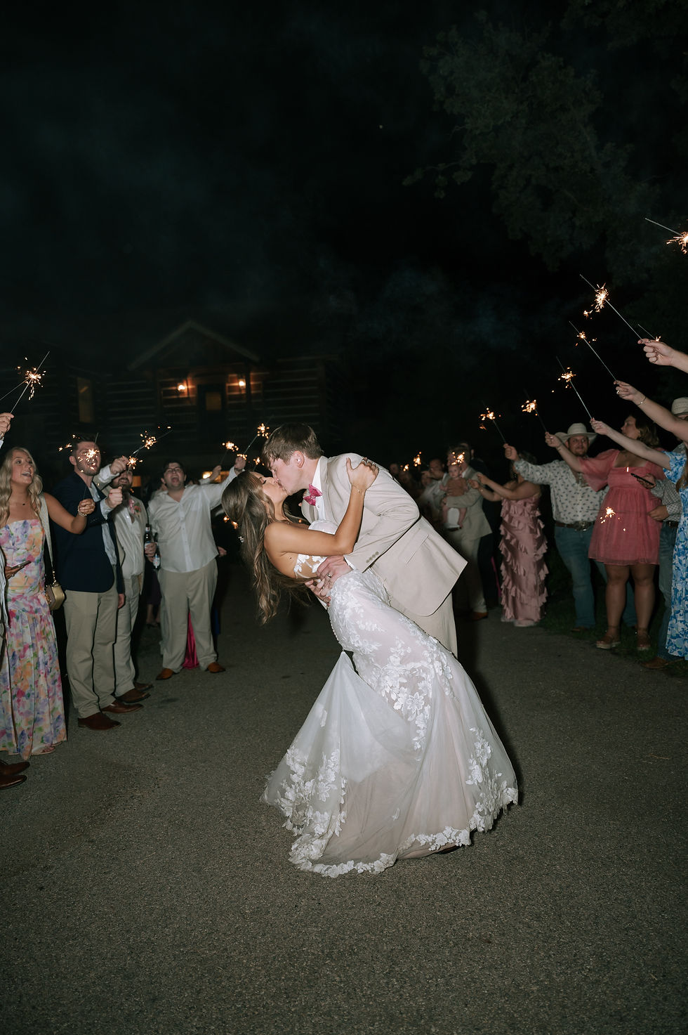 Bride and groom kiss surrounded by guests holding sparklers at night. Bride in lace dress, groom in beige suit, romantic celebration mood.