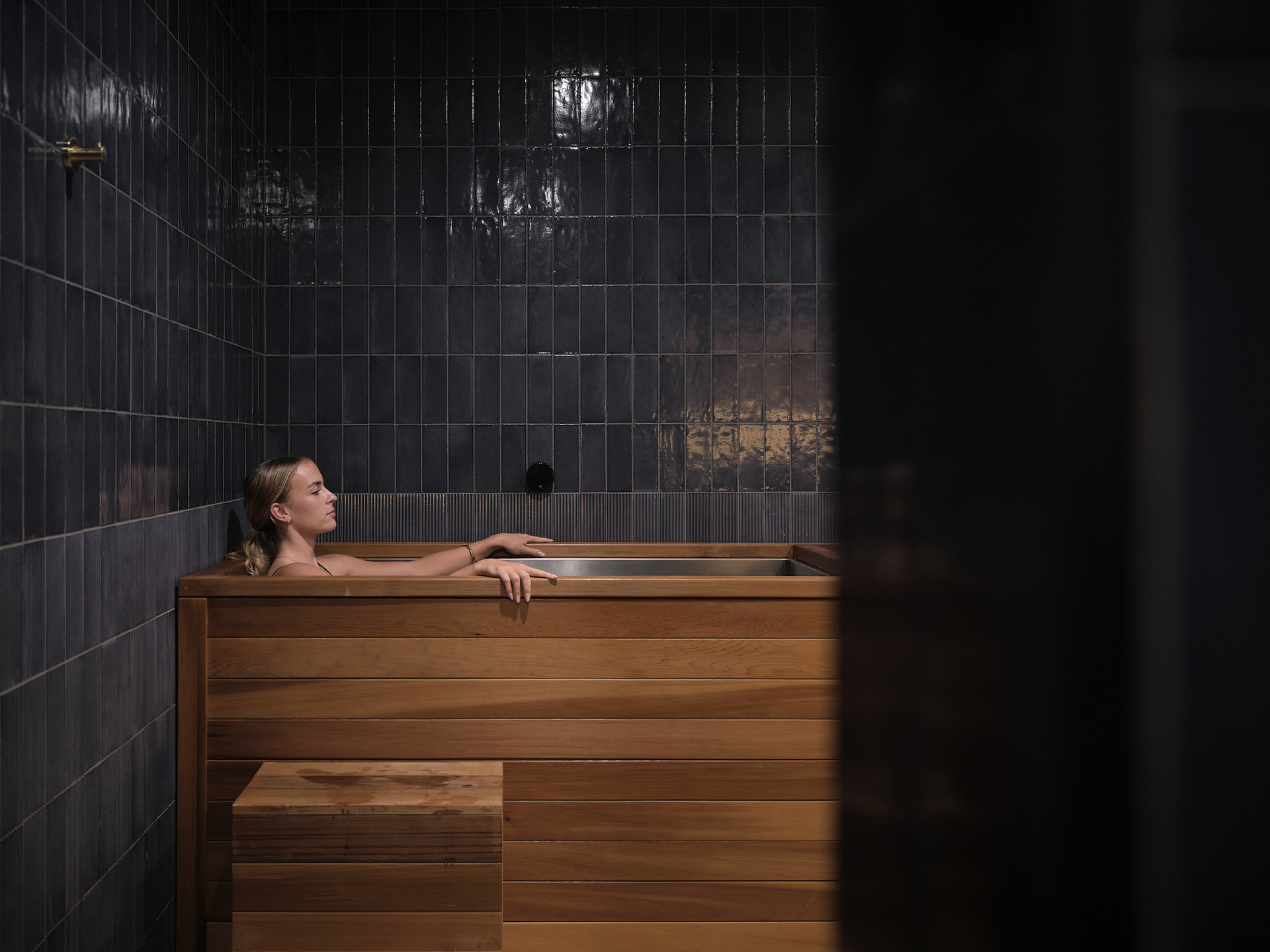 A woman resting in the cedar cold plunge against navy tiles at Boon Wellness