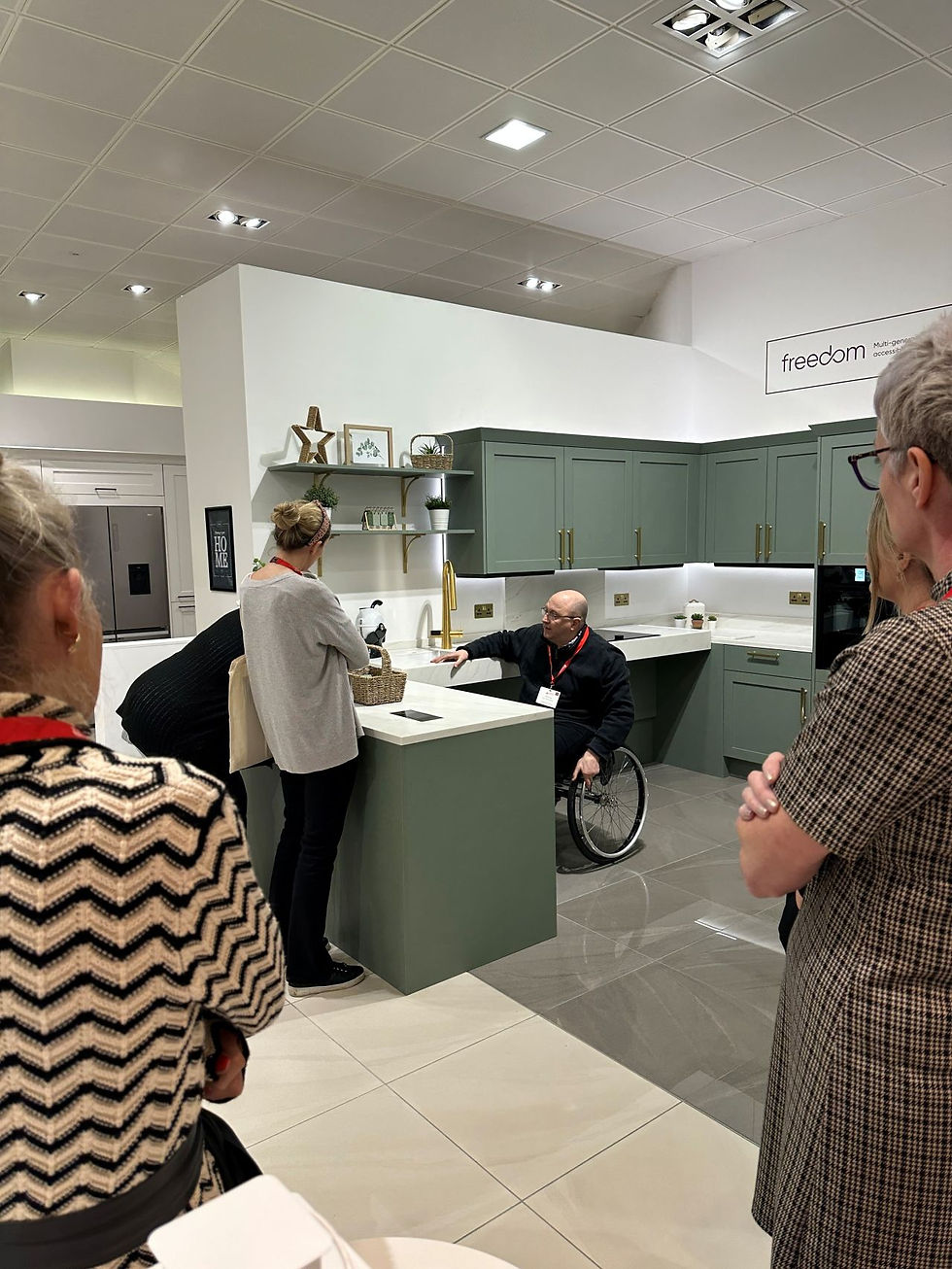 Image shows Adam Thomas demonstrating the straight rising fall sink and hob in a u-shaped soft green wheelchair accessible kitchen. People are standing and watching with their backs to camera. 