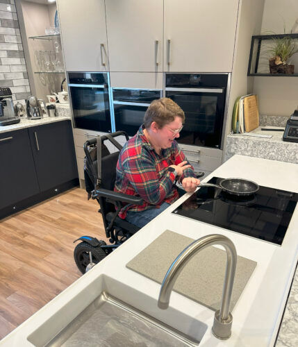 Image shows a seated wheelchair user with a red checked shirt in a u-shaped wheelchair accessible kitchen with blue base units to left, tall pale stone coloured units to centre of image with integrated accessible ovens, and rise and fall cooking and food prep zone on a solid surface worktop to right of image.