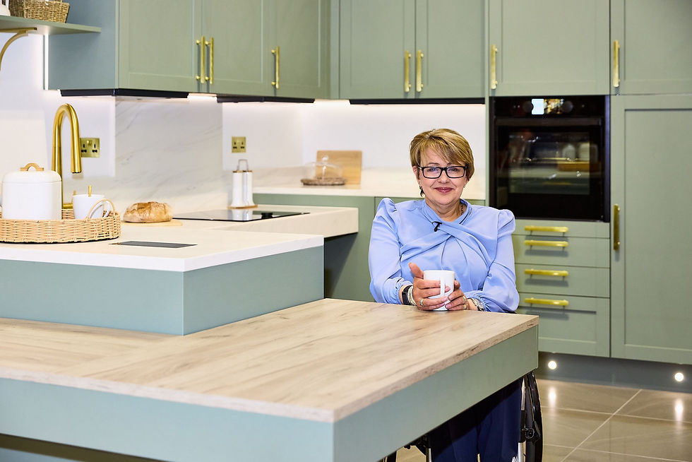 Baroness Grey-Thompson enjoys a coffee, seated at a breakfast bar in a soft green Freedom Kitchen with stone effect worksurface, and off white worksurfaces elsewhere in the kitchen.