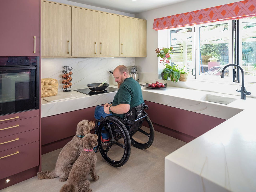 Image shows male client, seated in wheelchair to centre, with two dogs  seated next to him. Large u-shaped kitchen with tall unit and integrated oven to near left in soft merlot red colour, large l-shaped rise and fall hob and sink zones, with contrasting pale, textured wood cabinets over, and, and large solid surface peninsula to near right.