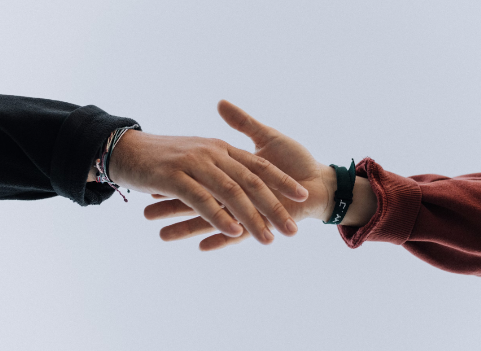 Two hands reaching to shake against a light blue background. One wears a red sleeve, the other black with bracelets.