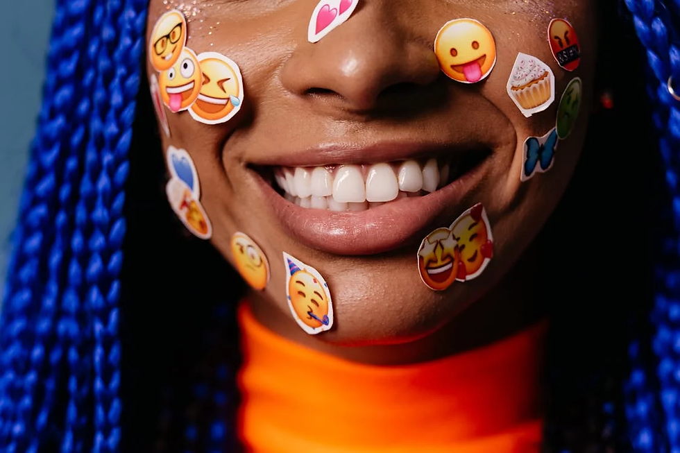 Smiling person with blue braids, wearing bright orange. Emoji stickers cover their face, creating a playful and joyful mood.