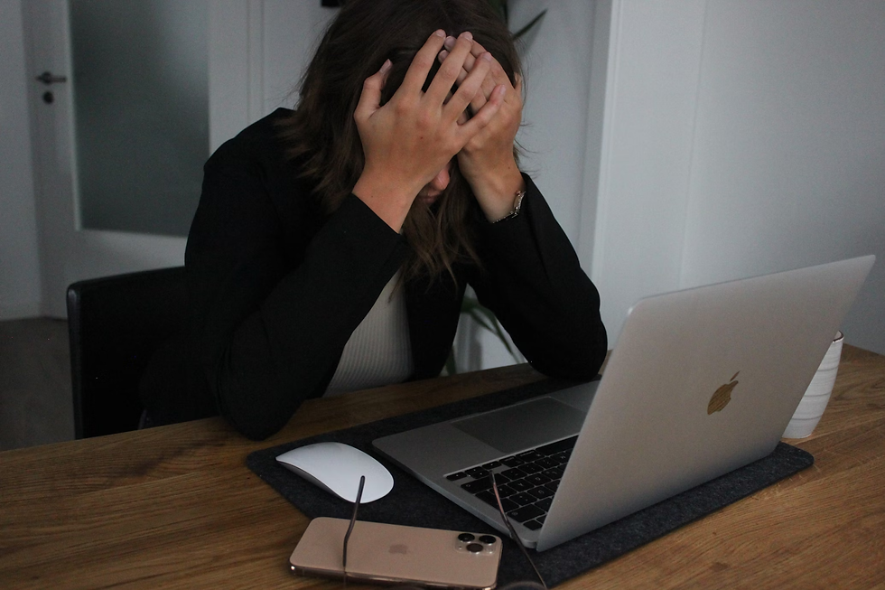 Person sitting at a desk, head in hands, looking stressed. Open laptop and phone on the wooden table. Minimalistic room background.