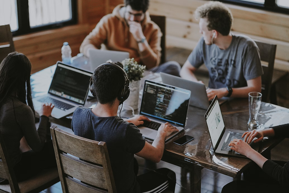 Five people collaborate with laptops around a wooden table in a cozy room. One wears headphones, another a brown hoodie. Casual, focused mood.