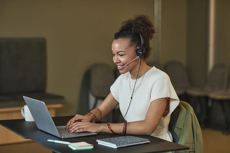 Smiling woman with a headset types on a laptop at a desk. She wears a white top; a notebook and pen are nearby. Office chairs in background.