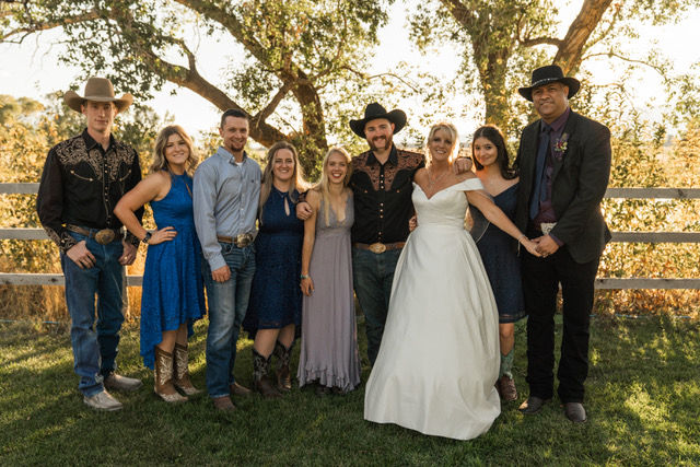 Wedding group photo outdoors. Bride in white dress, groom and guests in cowboy hats. Smiling, standing on grass, trees in background.