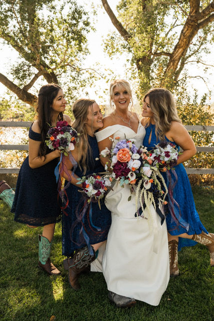 Bride in white and three bridesmaids in blue dresses with cowboy boots pose joyfully, holding bouquets, outdoors near a wooden fence.