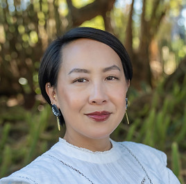A person with short dark hair, wearing a white shirt, smiling gently at the camera, standing before a forest background.