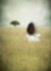 Woman with long dark flowing hair blowing in the wind stands in a field of tall grass looking out towards the horizion where she sees a tree standing tall and majestic.
