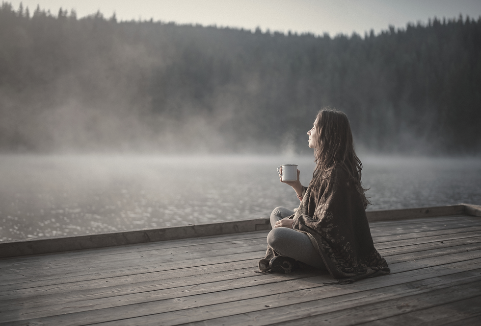 Woman sits on a wooden dock by a misty lake, holding a mug and gazing at the forested shore in a calm, reflective mood.