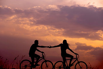 silhoutted image of a couple riding bikes while reaching out to hold each other's hand. There is a sunset in the background