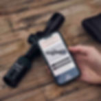 Ein Zielfernrohr liegt auf einem Holztisch neben einem Smartphone, das eine Bedienungsanleitung für das Zielfernrohr anzeigt.