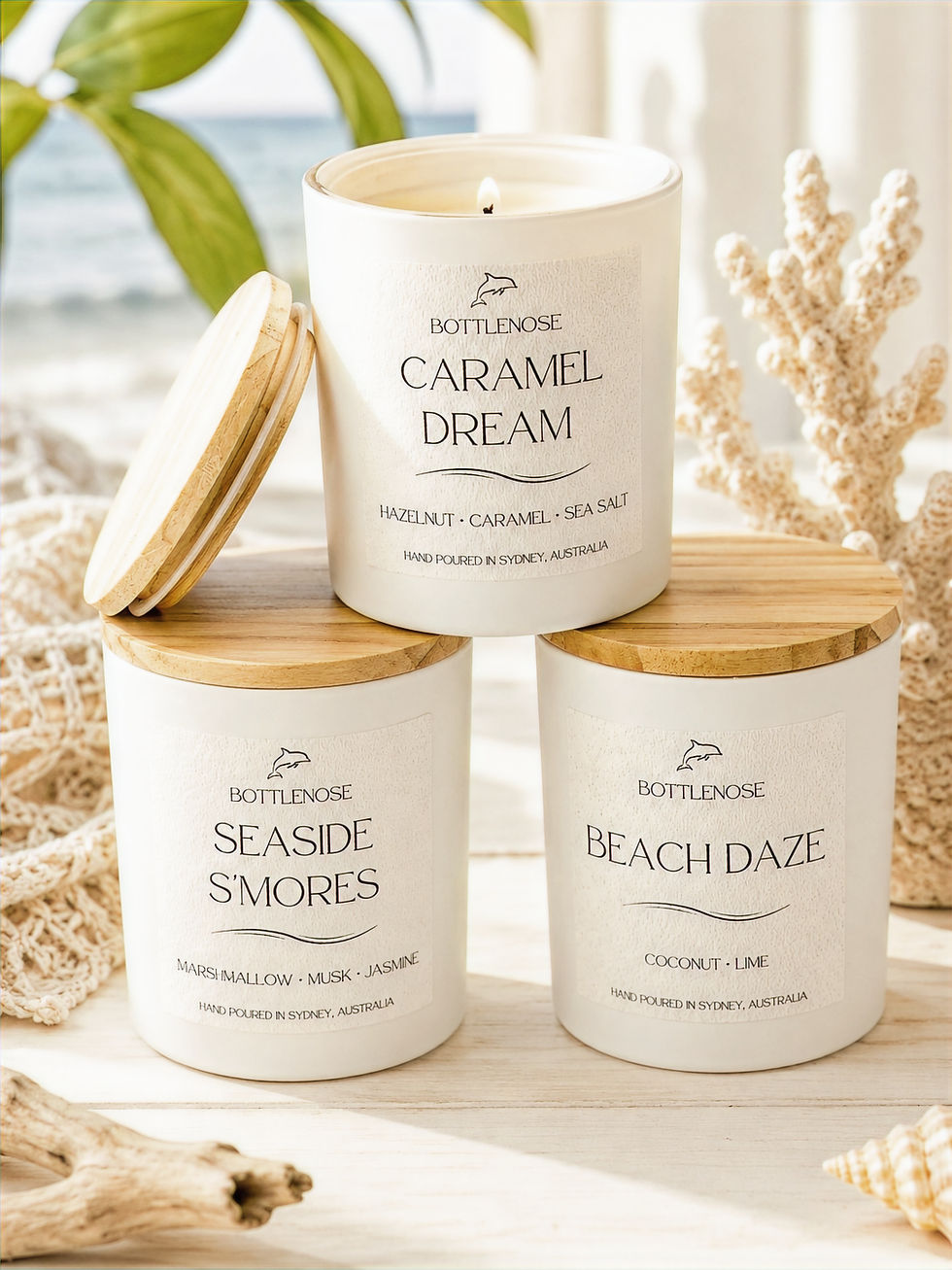 Three white Bottlenose candles on a wood table with beach shells; labels read Caramel Dream, Seaside S'mores, and Beach Daze.