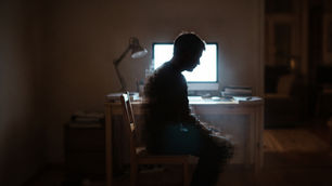 Silhueta de uma pessoa em um quarto escuro sentada em frente a um monitor de computador brilhante, ilustrando o isolamento e o vício em pornografia discutidos no artigo.