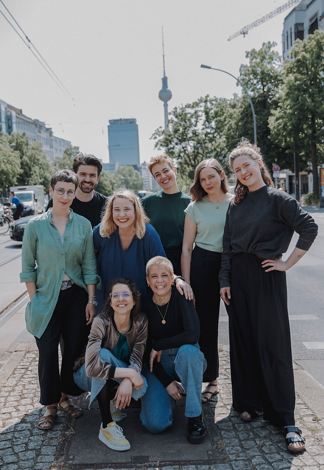 Gruppenbild der Lehrer*innen von SDG an der Tramhaltestelle vor der Schule. Lachend, im Hintergrund der Berliner Fernsehturm