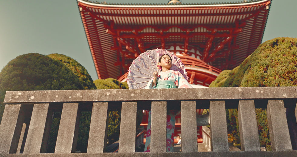 Woman in kimono holds parasol, standing on stone balcony. Red Japanese temple and green trees in background. Serene mood.