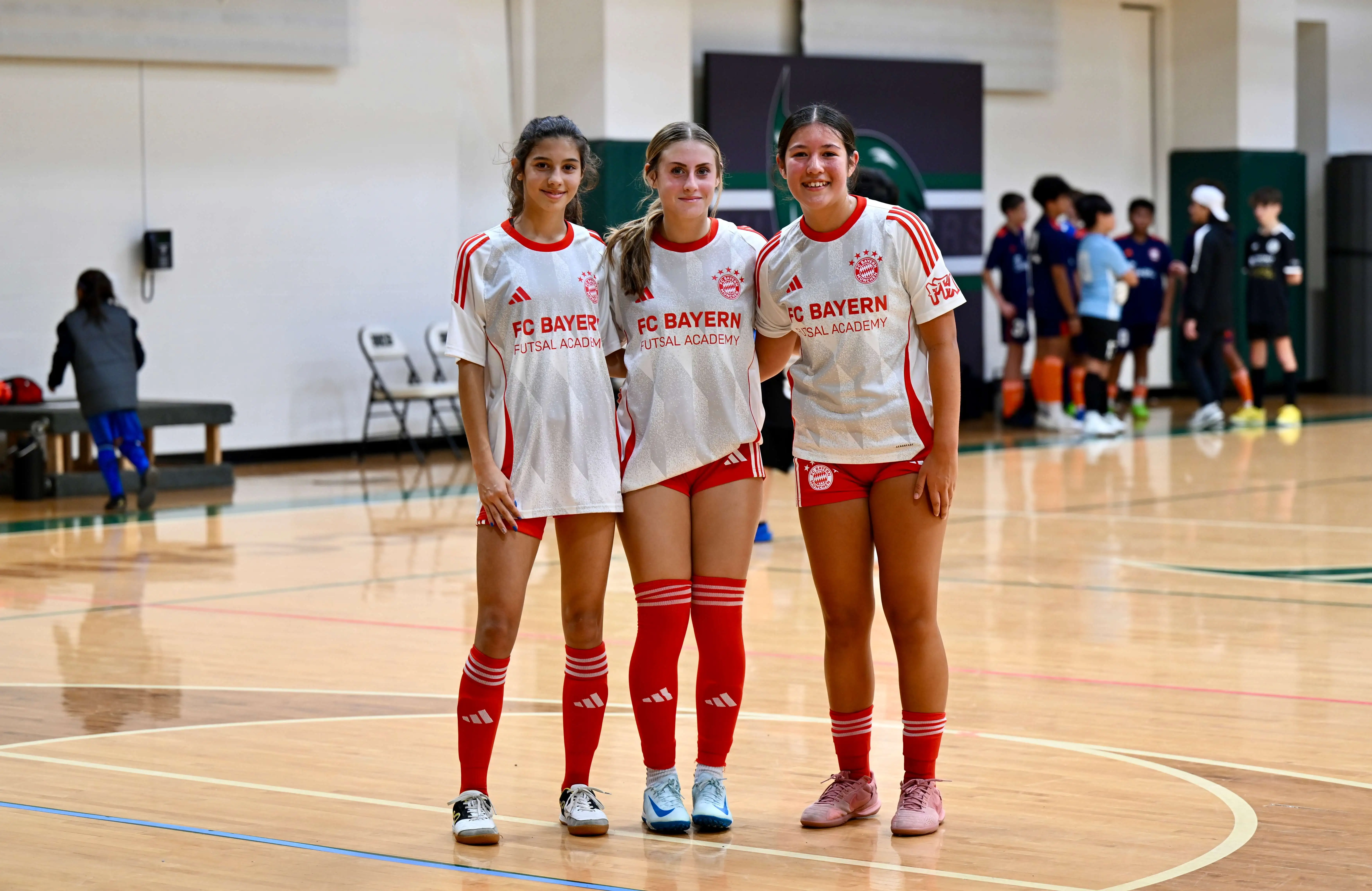 Three girls standing and posing on a futsal court while wearing their FC Bayern Futsal kits