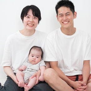 白い背景で、笑顔の夫婦が赤ちゃんを膝に抱いた家族写真。白Tシャツ姿で、赤ちゃんはグレーの服。