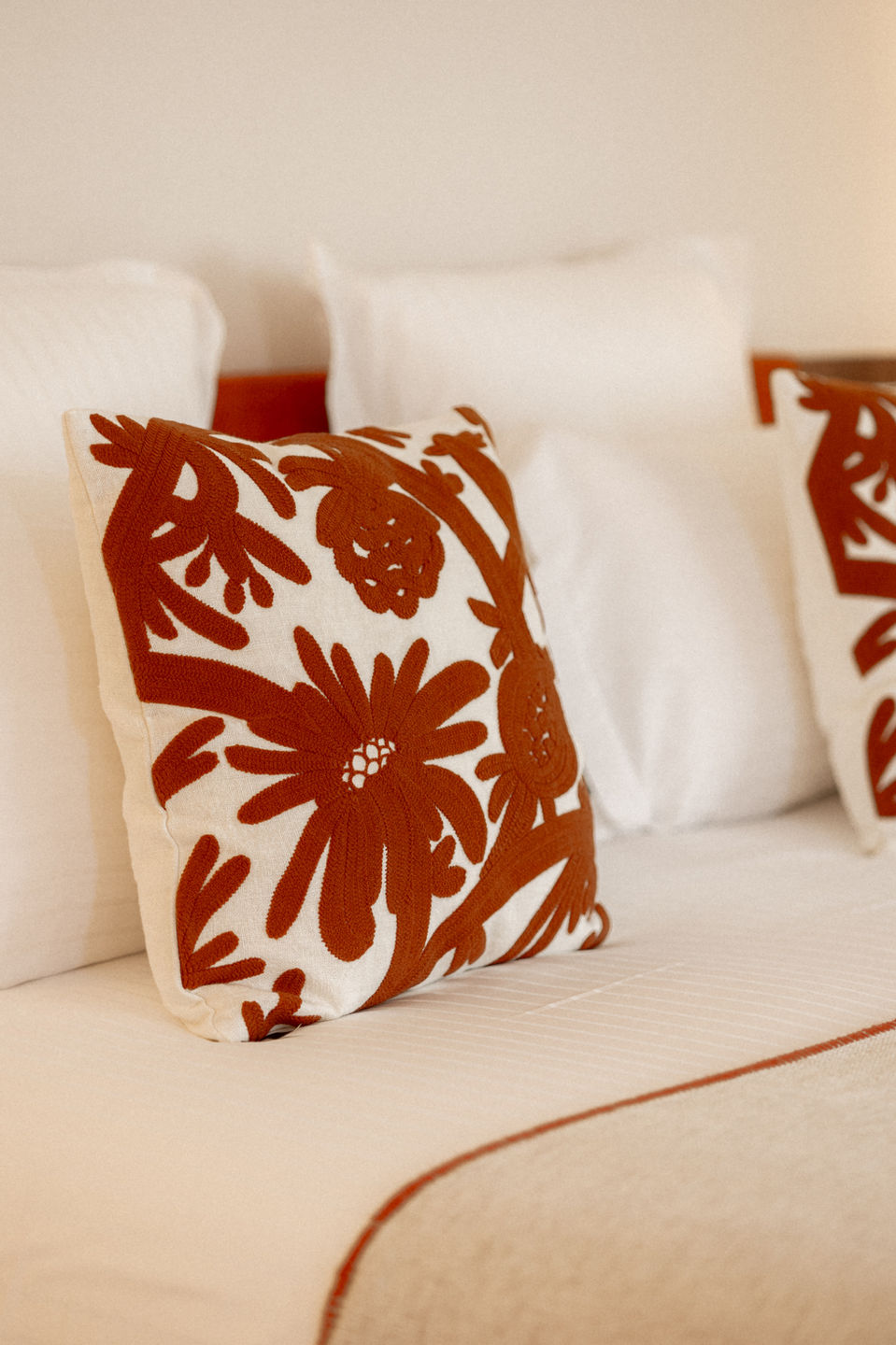 Coussin brodé terracotta à motifs floraux sur un lit avec linge blanc et détails en teintes naturelles.