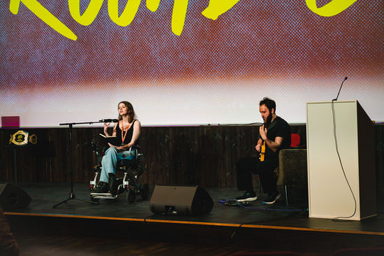 Photograph of Georgia Francis performing on stage in Loud Poets' Paisley Slam Heat at the Paisley Town Hall as part of Paisley Book Festival. She is accompanied musically by musician/photographer, Jack Hinks, who is playing guitar.