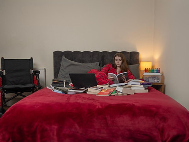 Photo of Georgia Francis in pyjamas, sitting on her bed surrounded by books. Her wheelchair is beside the bed.