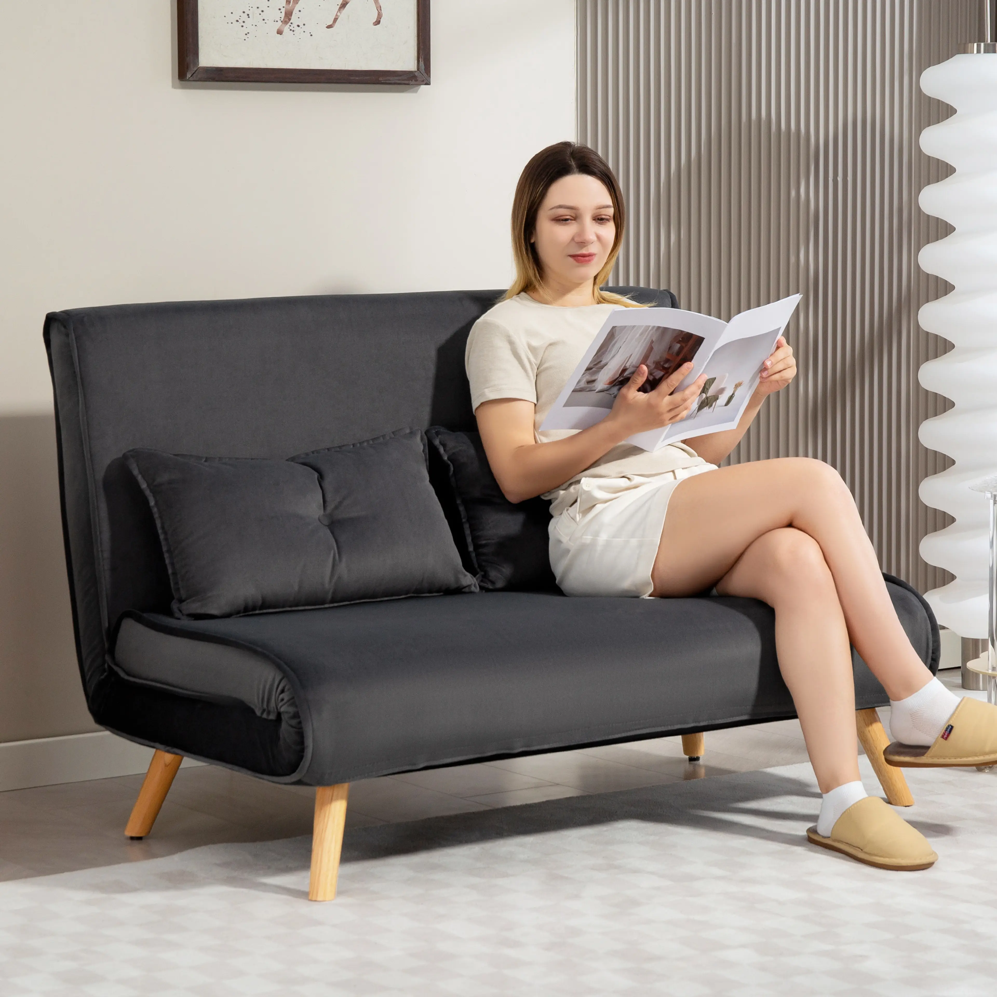 Mörkgrå bäddsoffa i soffläge med en kvinna som läser en tidning i ett rum med ribbad vägg och golvlampa