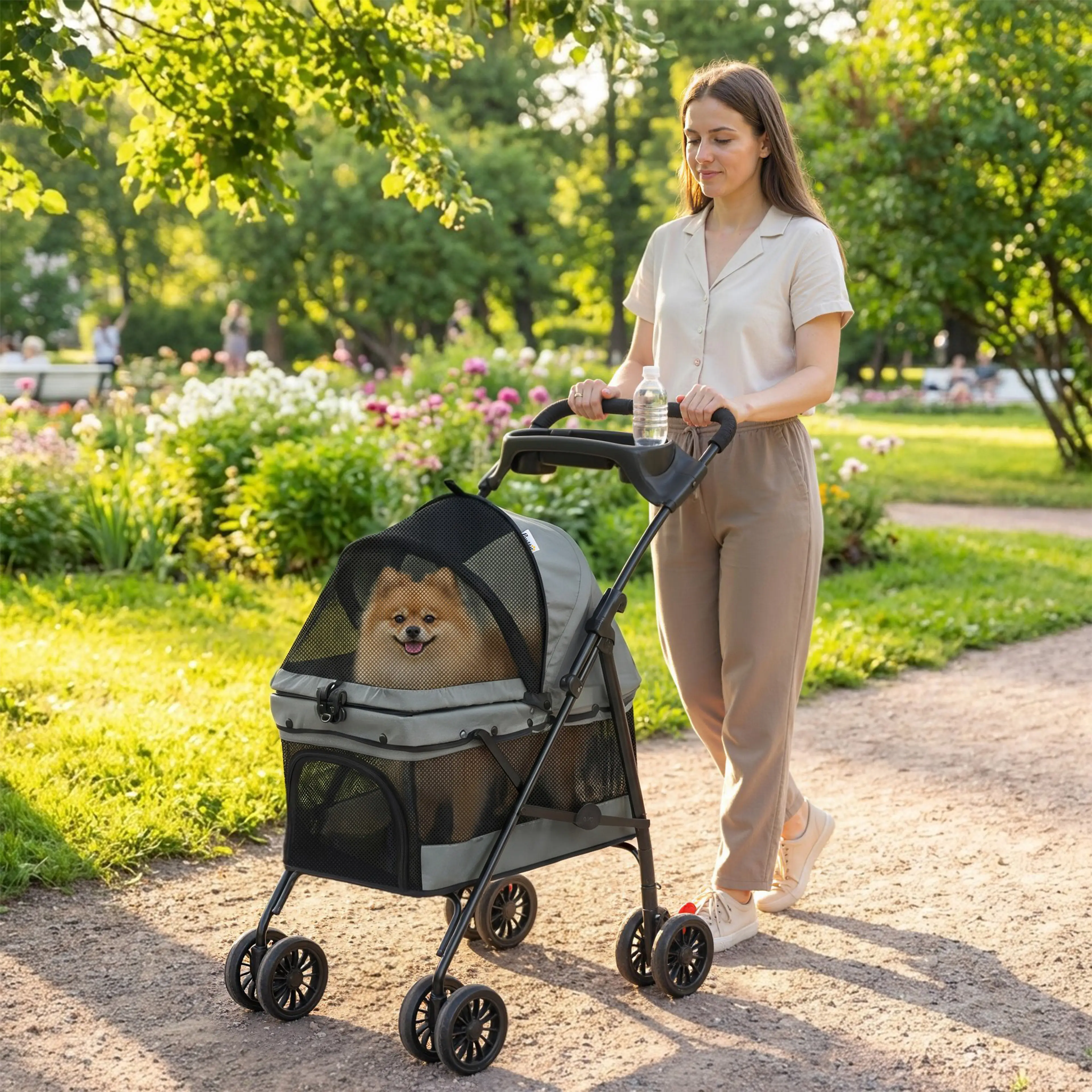 Kvinna går med den grå hundvagnen längs en grusgång bland blommor, med en liten hund som tittar ut genom nätfönstret och en vattenflaska i handtaget