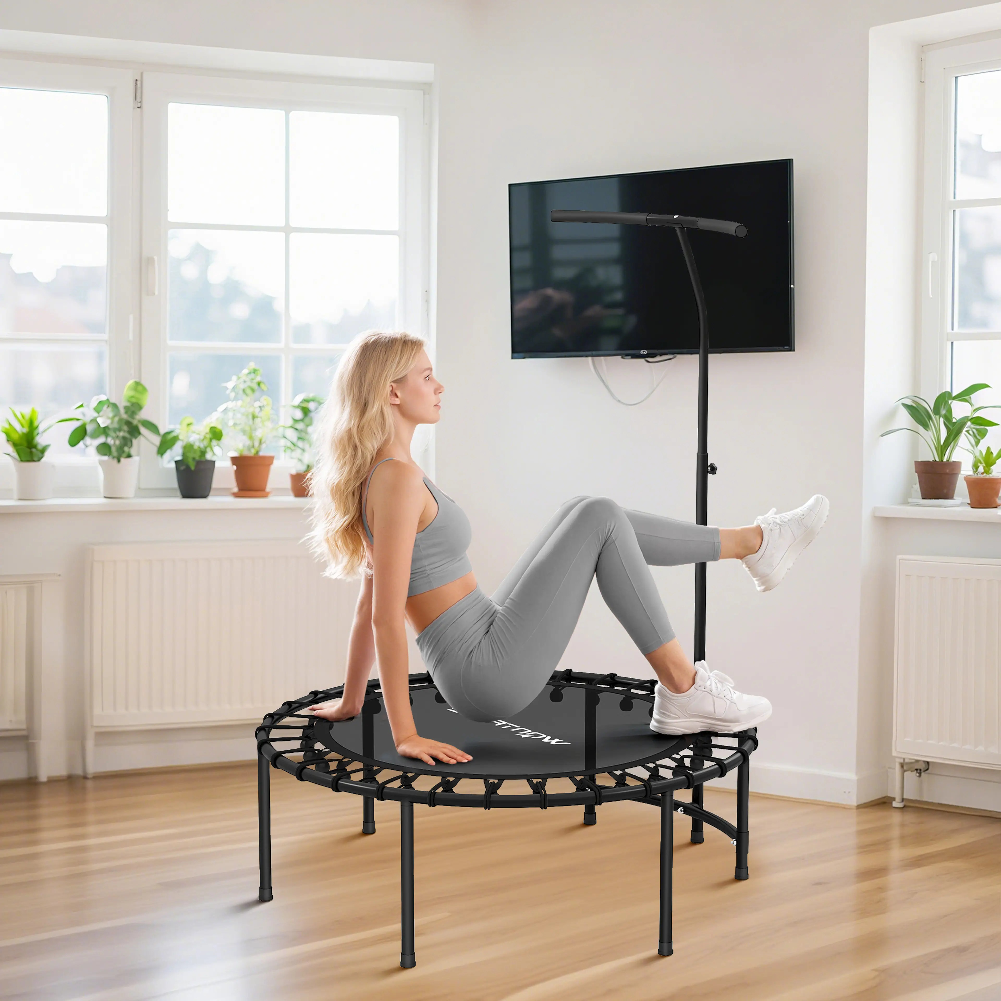 En kvinna i grå träningskläder sitter på studsytan och lutar sig bakåt på händerna med böjda ben, i ett ljust rum med krukväxter, radiator och tv på väggen