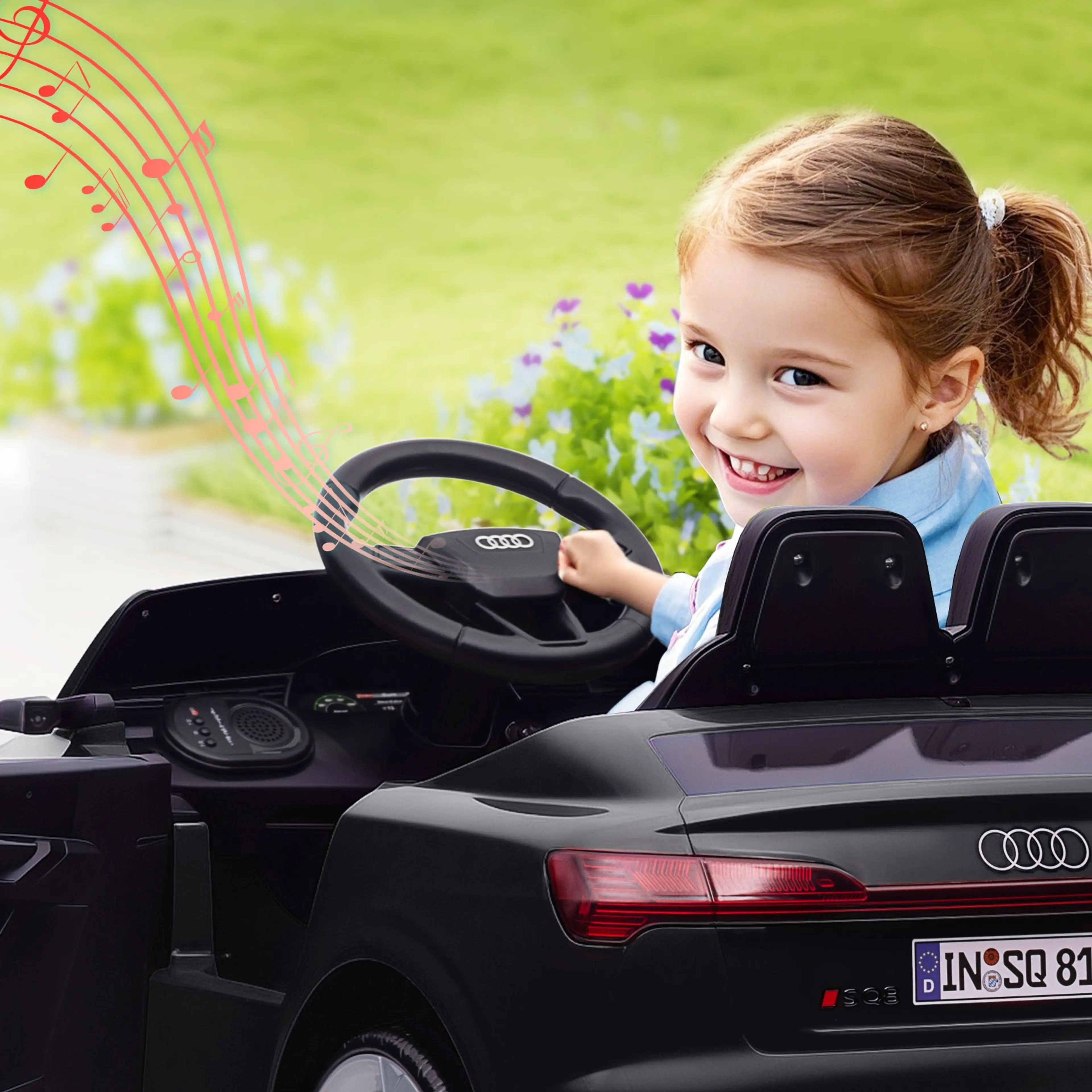 Leende flicka som håller i ratten i den svarta elbilen för barn, utomhus på gräs.