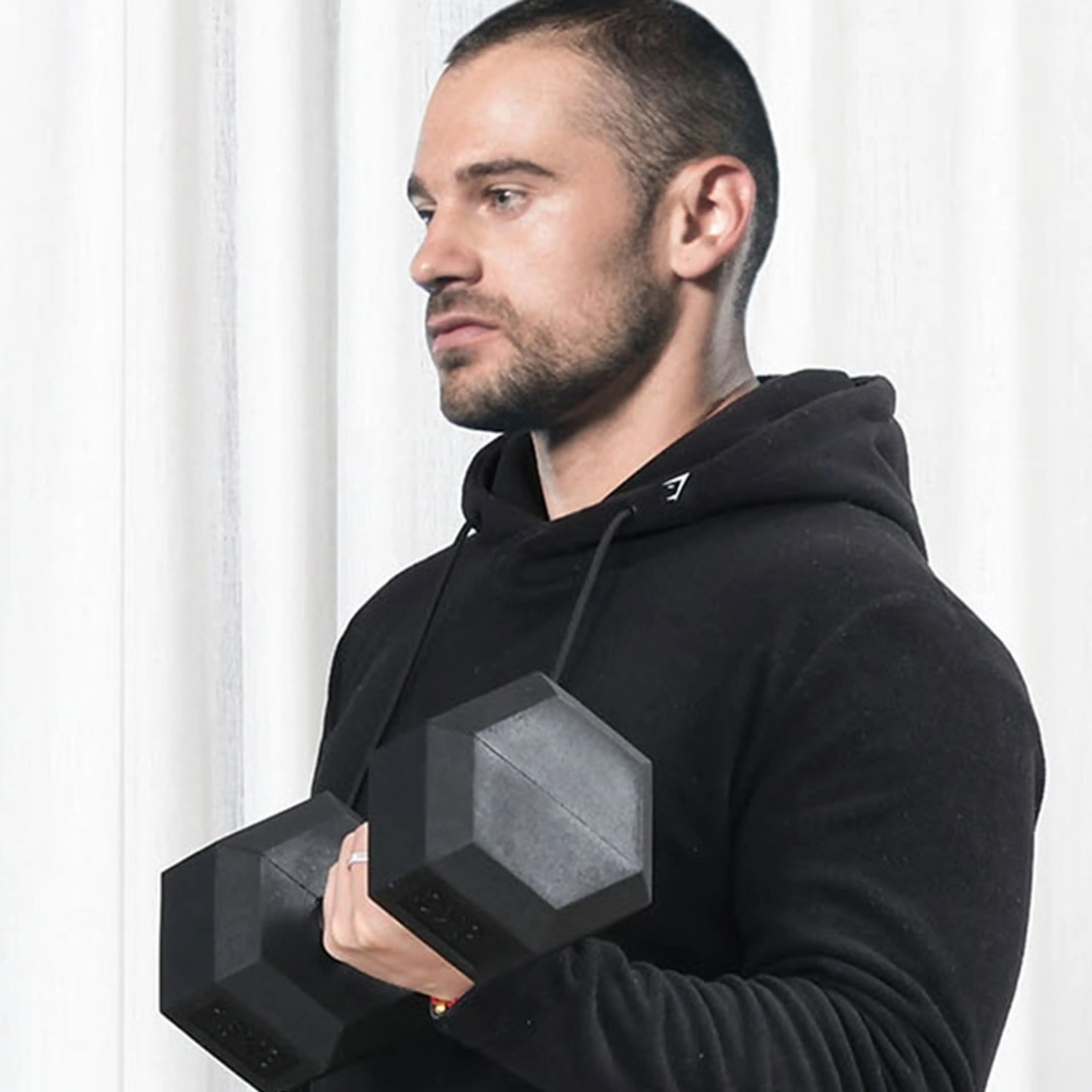 Man i svart hoodie håller hexhanteln 15 kg mot bröstet framför ett ljust draperi.