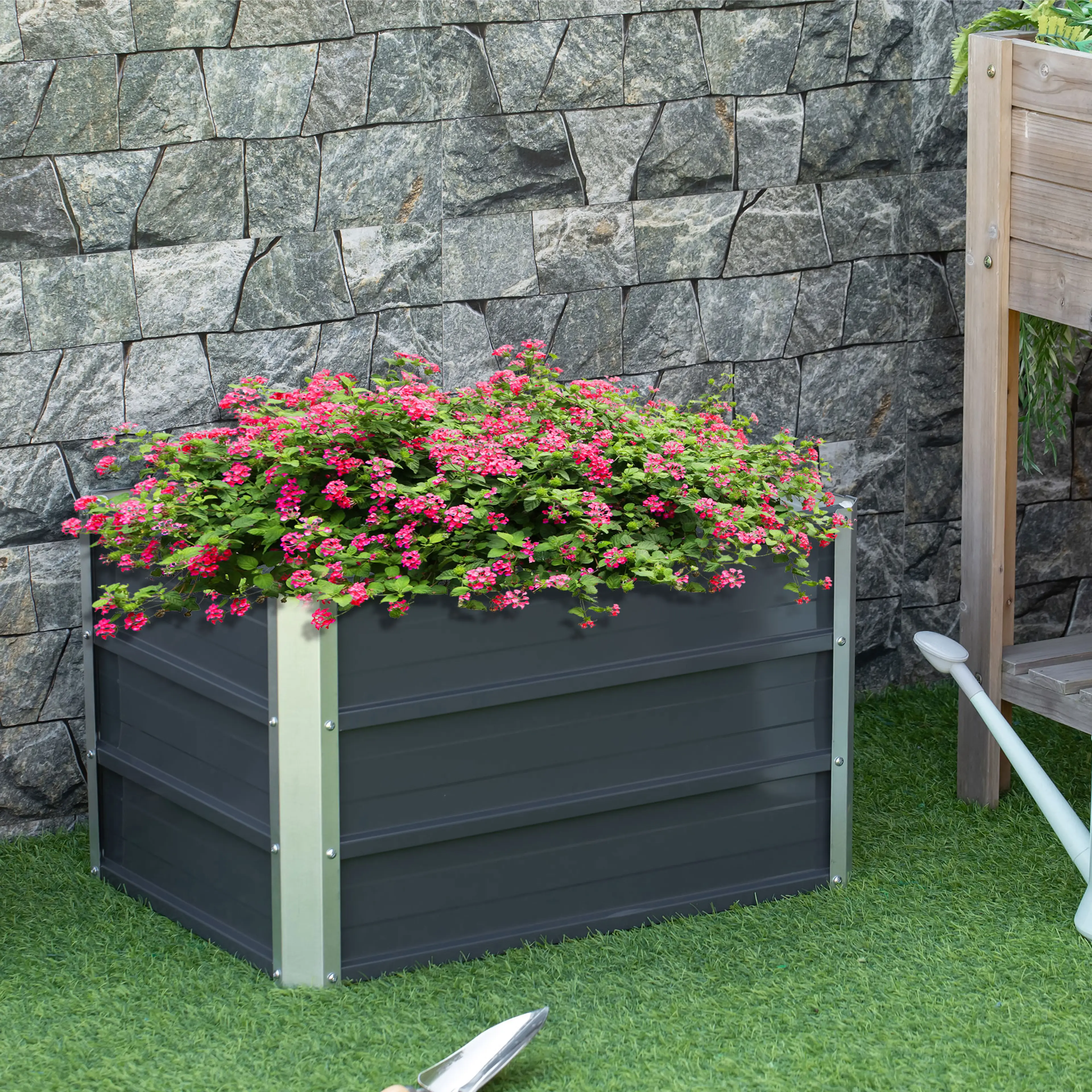 Odlingslådan fylld med rosa blommor framför en stenmur på gräsmattan