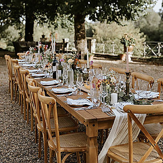 Long rustic wooden banquet table with cross-back chairs set for an outdoor corporate reception under mature trees at Château Destinée, wildflower centrepieces with candles and linen in dappled garden light, Dordogne France