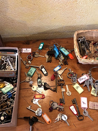 Collection of antique skeleton keys and modern room keys with handwritten French labels spread across a rustic wooden table at Château Destinée, sorted for guest room preparation