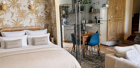 Grenache apartment: double bed with rattan headboard against golden palm wallpaper, with a dining area and kitchen with a retro fridge behind