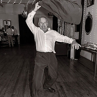 Guest tossing jacket in celebration on barn dance floor in black and white, joyful reception moment with string lights and rustic wooden beams at French château