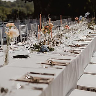 Long outdoor wedding banquet table with white linen, tall blush taper candles, wildflower arrangements in soft blue and peach tones, crystal glassware and white Chiavari chairs under open sky at Château Destinée Dordogne France