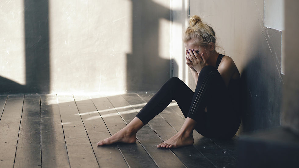 A person sits on a dark wooden floor against a wall, head in hands, appearing upset. Soft light creates shadows on the floor and wall.