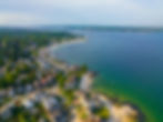 Aerial view of a coastal Connecticut town with houses, a curved shoreline, and a lighthouse. The sea is calm with shades of green and blue.